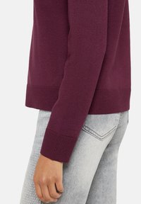 Bordowy sweter dziany z prążkowanymi mankietami i ściągaczem, w połączeniu z jasnoszarymi dżinsami. Tkanina wydaje się gładka, a krój przylegający do ciała.