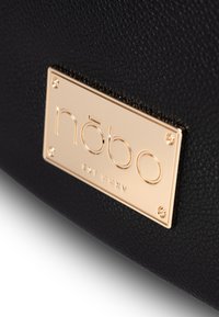 Schwarze strukturierte Handtasche mit einer goldenen Metall-Logo-Plakette mit eingraviertem Text und Eckakzenten. Nahaufnahme, die Materialien und Details betont.