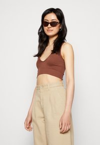 Top cropped castanho com textura e decote em V, combinado com calças beje de cintura alta e pregas. O modelo usa óculos de sol castanhos em forma de gato.