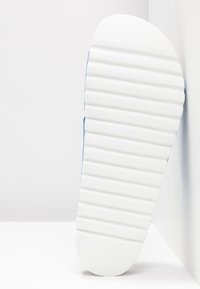 Semelle de chaussure en caoutchouc blanc avec des rainures texturées, positionnée sur un fond clair. Bout arrondi avec une touche de bleu le long du bord.