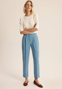 Jasnoniebieskie dopasowane spodnie z fałdami z przodu, połączone z białą teksturowaną bluzką i brązowymi płaskimi butami ozdobionymi złotymi akcentami.