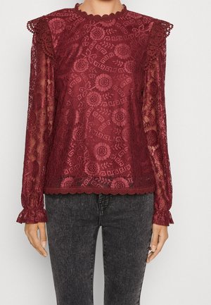 Kobieta w długim rękawie bordowej koronkowej bluzce z kwiatowymi wzorami i marszczonymi ramionami, zestawiona z czarnymi, spranymi dżinsami.