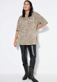 Overmaatse beige shirt met luipaardprint en korte mouwen, gecombineerd met zwarte nepleer leggings en zwarte cowboylaarzen met geperste details.