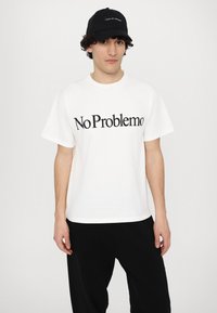 White cotton t-shirt featuring black "No Problemo" text. Accompanied by a black cap with white text. Model stands against a white background.