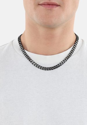 Metallkette mit schwarzem Finish, bestehend aus ineinandergreifenden Gliedern, liegt auf einem weißen, strukturierten Hemd in der Nähe des Ausschnitts.
