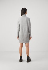 Grijze gebreide jurk met een hoge kraag, lange mouwen en een lengte tot halverwege het bovenbeen, gecombineerd met zwarte veterschoenen. Glad materiaal en een relaxte pasvorm.