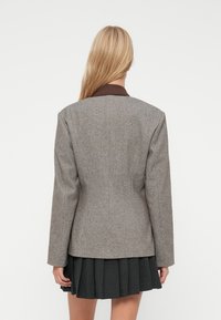 Blazer gris de espiga con diseño estructurado, solapas de muesca y mangas largas, llevado sobre una falda negra con pliegues. Tejido texturizado.