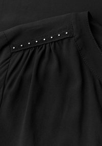 Svart blus med texturerad axel prydd med dekorativa silvernitar och veckad tygdetalj. Slät tyg med enkel design.