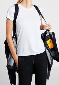 Personne portant une chemise blanche à manches courtes et un pantalon noir texturé, ajustant une veste noire et grise avec une étiquette jaune visible à l'intérieur.