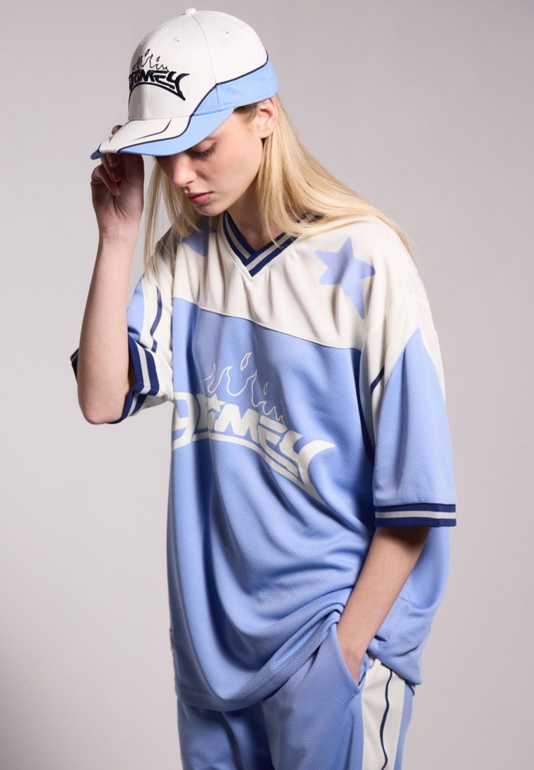 Lichtblauwe oversized jersey met witte accenten en blauwe strepen, voorzien van een vlamlogo. Witte pet met blauw en zwart detail.