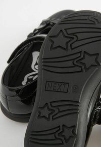 Zapatos de charol negros con una correa de velcro, que presentan una suela texturizada con patrones de estrellas y la marca "NEXT" grabada en el talón.