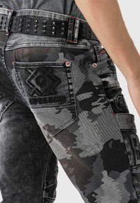 Jean camouflage noir et gris avec un tissu texturé, des coutures contrastées, une ceinture noire cloutée et des détails en relief sur la poche arrière.