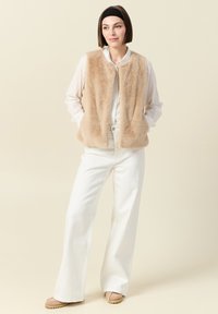 Beige fuskpäls väst med rund halsringning och framfickor, parat med en vit skjorta och vida vita byxor. Ljus bakgrund.