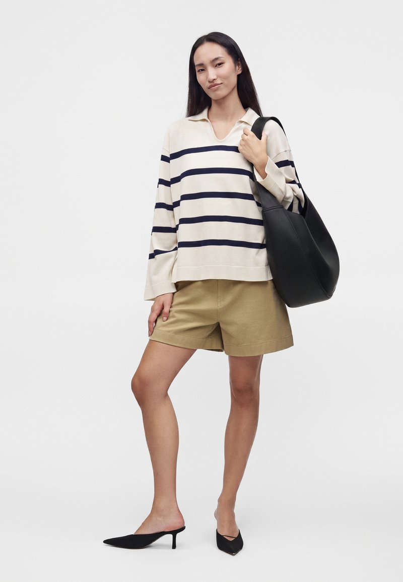 Femme portant une chemise à manches longues rayée crème et marine, un short beige, des talons noirs, tenant un grand sac à bandoulière noir.
