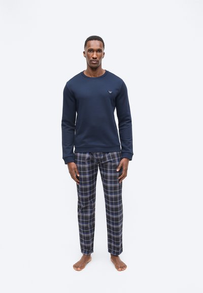 Άντρας που στέκεται ξυπόλητος, φορώντας μπλε σκούρο πουλόβερ και μπλε καρό πιτζάμες, με φόντο λευκό και απλό.