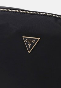 Zwart nylon etui met een gouden ritssluiting. Heeft een driehoekig metalen logo in goud en zwart met de tekst "GUESS USA". Glad oppervlak.