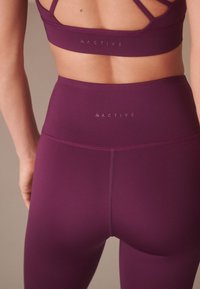 Violettes Sportoutfit bestehend aus einem Sport-BH mit dünnen Trägern und hoch taillierten Leggings. Glatte Textur mit minimalen Nähten und Logodetail.