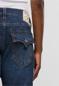 Temno modri džins hlače z blagovno znamko, poševnimi zadnjimi žepi s preklopom in kontrastnimi oranžnimi šivi.