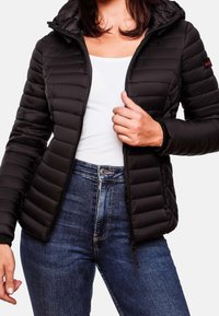 Žena oblečená v černé prošívané bundě přes bílé tričko a modré džíny, zapínající bundu.