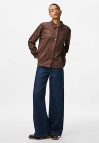 Brun mockaskjorta med krage och knäppning fram, tillsammans med vida mörk denimjeans. Detaljer inkluderar stora fickor och robusta skor.