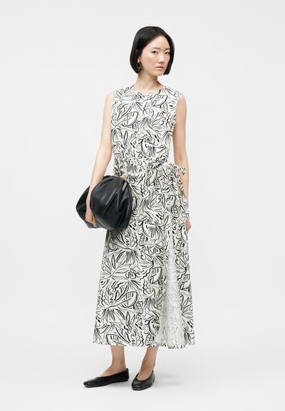 Γυναίκα με ίσια μαύρα μαλλιά φοράει λευκό αμάνικο φόρεμα με μαύρο αφηρημένο σχέδιο φύλλων, κρατάει μια μεγάλη μαύρη τσάντα και μαύρα επίπεδα παπούτσια.