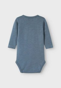 Langsleeve babypakje van zacht, lichtblauw materiaal met een subtiele textuur. Het heeft een ronde halslijn en een drukknoopsluiting aan de onderkant.