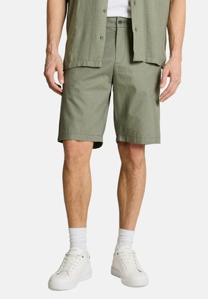 Mann trägt olivgrüne, geknöpfte Shorts, kombiniert mit weißen Sneakers und weißen Kniestrümpfen, steht vor einem schlichten Hintergrund.