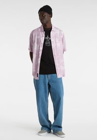 Camisa de manga corta rosa con estampado sobre una camiseta gráfica negra, combinada con vaqueros azules holgados y zapatillas de cuadros sin cordones. Se lleva un gorro negro.