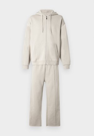 Sweatshirt bege com fecho de correr, bolsos canguru e cordões, combinado com calças de ganga ajustadas com costuras laterais e textura suave.