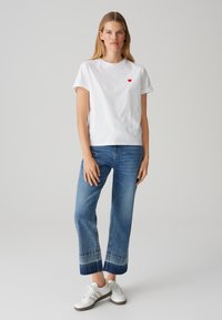 T-shirt bianca in cotone con logo a cuore rosso, abbinata a jeans blu cropped con orlo sbiadito e scarpe atletiche bianche.