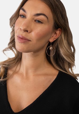 Frau mit hellbraunem welligem Haar, die große goldene Creolen mit einem Sonnenmotiv und ein schwarzes V-Ausschnitt-Top trägt, schaut zur Seite.