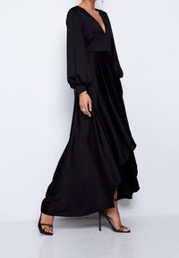 Kvinna klädd i en svart långärmad maxiklänning med V-ringning och asymmetrisk fåll, med svarta snörda högklackade sandaler, stående mot en vit vägg.