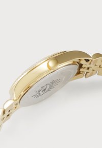 Montre-bracelet dorée avec un boîtier rond, une lunette texturée et un cadran argenté réfléchissant avec un motif solaire. Bracelet en maillons métalliques.