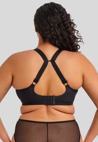 Vrouw in een zwarte sportbeha met racerback en zwarte mesh high-waisted onderbroek, gezien vanuit de rug tegen een eenvoudige grijze achtergrond.