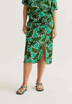 Mujer con falda midi verde con estampado de hojas tropicales y abertura frontal, combinada con un top a juego y sandalias de tiras verdes oscuras.