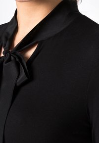 Svart topp med en satinnekband vid kragen, en nyckelhålsutskärning och mjuk, slät tygtextur. Nära bild av detaljerna vid halsringningen.