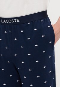 Navyblauwe broek met een zachte textuur, met een patroon van witte Lacoste-logo's. Elastische tailleband met contrasterende details.