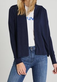 Marinblå öppen fram kardiagan gjord av ett mjukt tyg, med långa ärmar och en avslappnad passform, lager över en vit t-shirt med blå text.