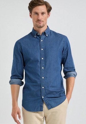 Man met lichtbruin haar die een lange mouwen blauwe denim shirt met opgerolde mouwen en beige broek draagt tegen een effen achtergrond.