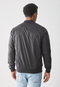 Sort bomberjakke med en glat finish, ribbet krave og manchetter, elastisk talje, set fra ryggen, parret med lyseblå jeans.