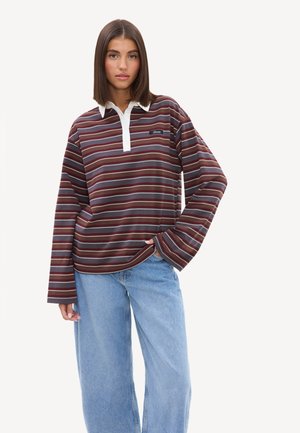 Jeune femme aux cheveux bruns raides portant un polo rayé à manches longues et un jean bleu clair ample, debout avec une main dans la poche.