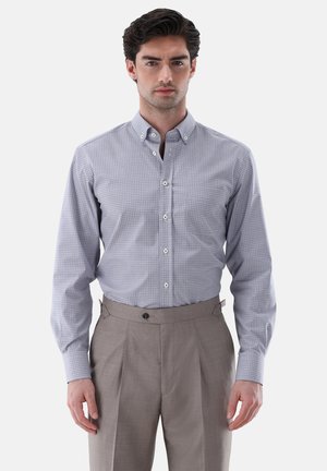 Mann trägt ein hellblau kariertes Hemd, das in hoch taillierte beige Hosen gesteckt ist, steht vor einem schlichten Hintergrund.