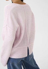 Pull en tricot rose clair avec une coupe décontractée, bords côtelés et couture centrale. Le dos présente une petite fente au-dessus de l'ourlet. Associé à un jean bleu.