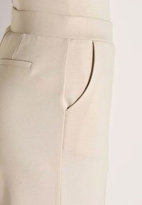 Close-up van de tailleband van beige broek en detail van de zijzak met een achterzak op een neutrale achtergrond.