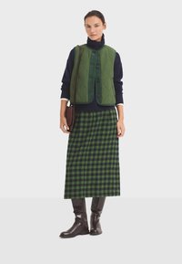 Chaleco acolchado verde sobre un suéter de cuello alto azul marino, combinado con una larga falda de cuadros verde y negra y botas marrones hasta la rodilla. Bolso marrón.
