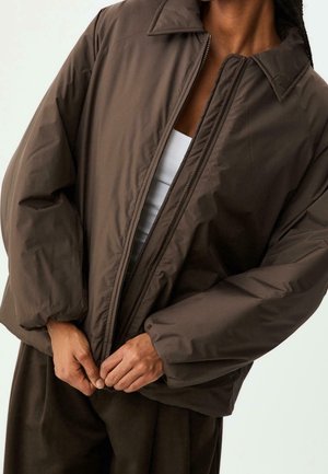 Personne portant une veste matelassée ample de couleur marron foncé avec une fermeture éclair, partiellement ouverte pour révéler un haut blanc en dessous.