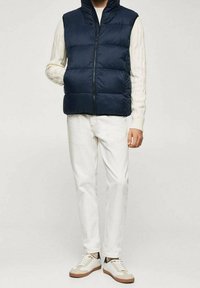 Tummansininen puffertakki, jossa on korkea kaulus, vetoketjukiinnitys ja tikattu muotoilu. Takki on päällä vaalean kreeminvärisen neuleen, ja se on yhdistetty valkoisiin housuihin ja lenkkareihin.