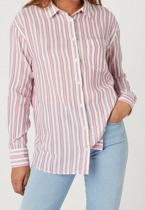 Nainen, jolla on päällään pitkähihainen, napitettava paita, jossa on vaaleanpunaisia, valkoisia ja harmaita pystysuoria raitoja, yhdistettynä vaaleansinisiin farkkuihin, käsi taskussa.