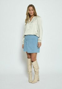 Blusa di satin beige chiaro con scollo a V, abbinata a una mini gonna in velluto a coste blu con bottoni sul davanti. Alti stivali beige con tacco.