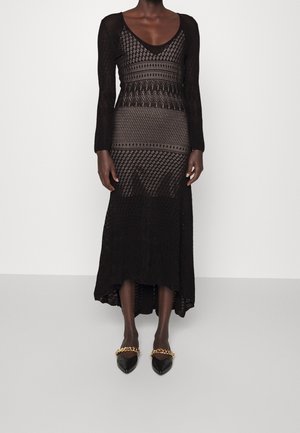 Kvinde iført en sort, langærmet, mønstret strikkjole med høj-lav kant og sorte spidse sko med gyldne kædedetaljer.